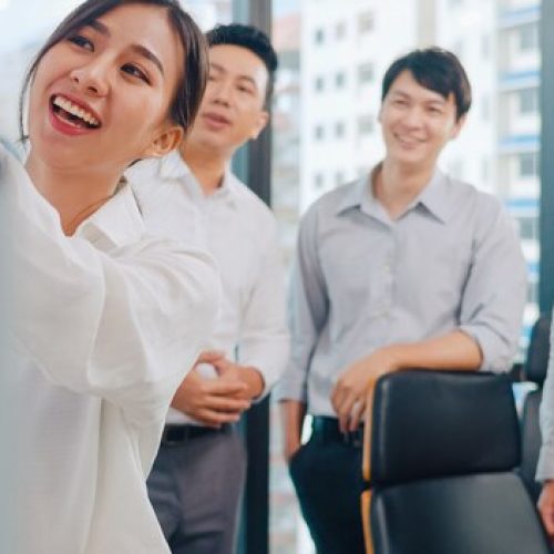 a woman pointing at a board with her colleagues in the background | ask2axcel